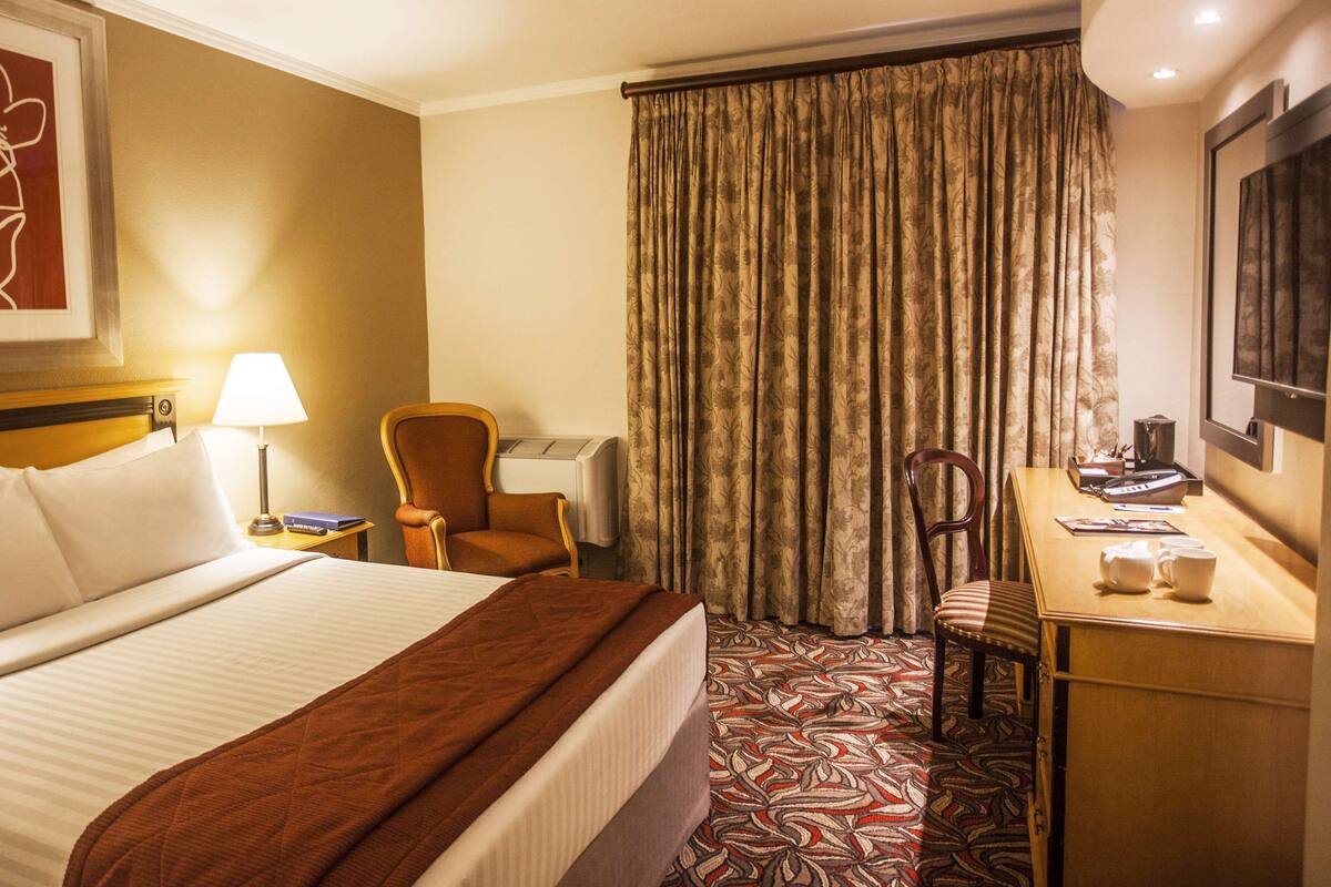 Een hotelkamer met een bed, bureau, stoel, lamp, koffiezetapparaat en een groot raam met gordijnen.