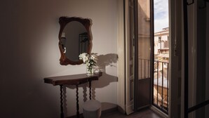 A room with a wooden console table, a mirror, a small stool, and a window with a view of buildings. - B&B Università (Catania)