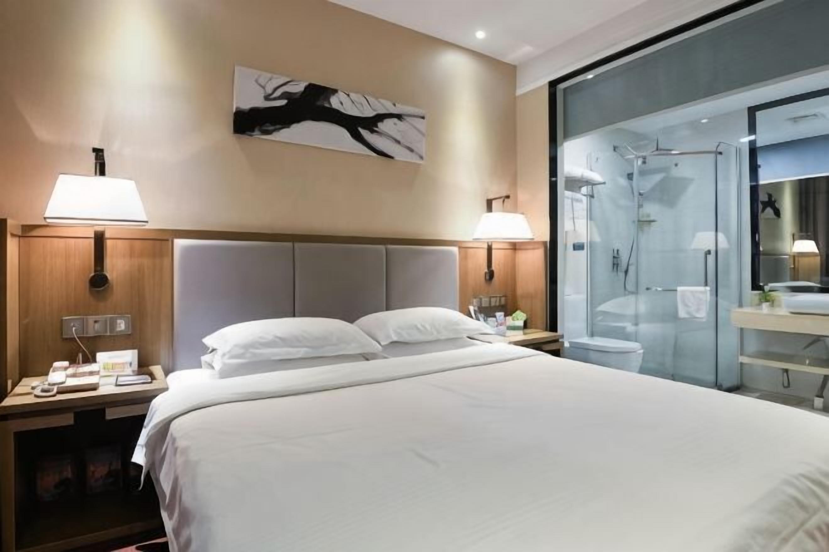 A hotel room with a large bed, two bedside lamps, a nightstand, a wall-mounted artwork, and a glass shower enclosure.