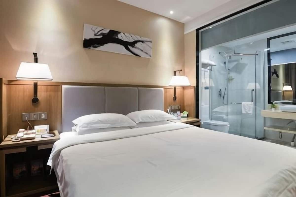 Habitación de hotel con una cama grande, dos lámparas de noche, una mesita de noche, una obra de arte colgada en la pared y un box de ducha de vidrio.
