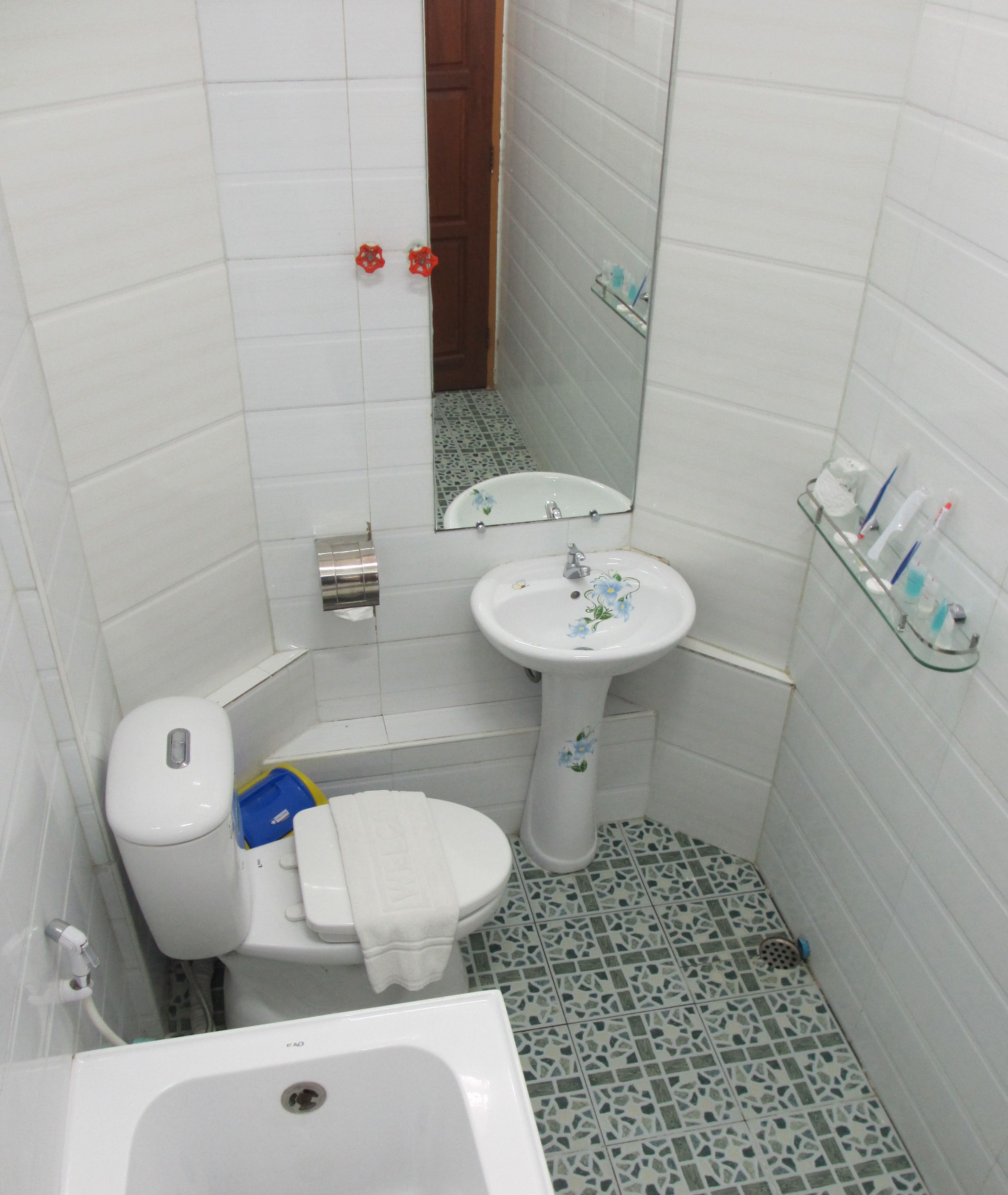 Um banheiro pequeno com vaso sanitário, pia e espelho. O chão possui um padrão de mosaico.