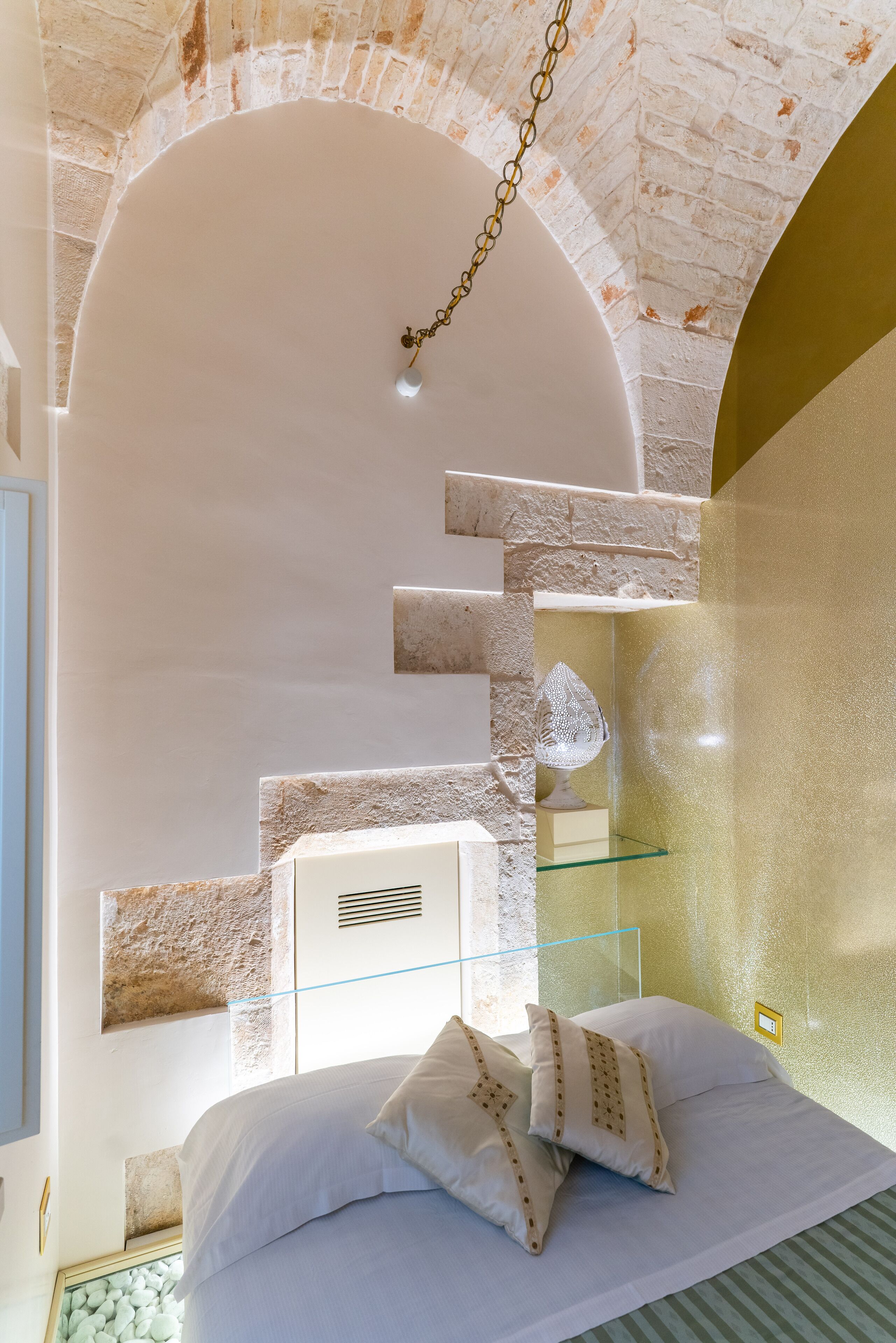 Un dormitorio con cama blanca, almohadones decorativos, estante de vidrio y pared de ladrillo.