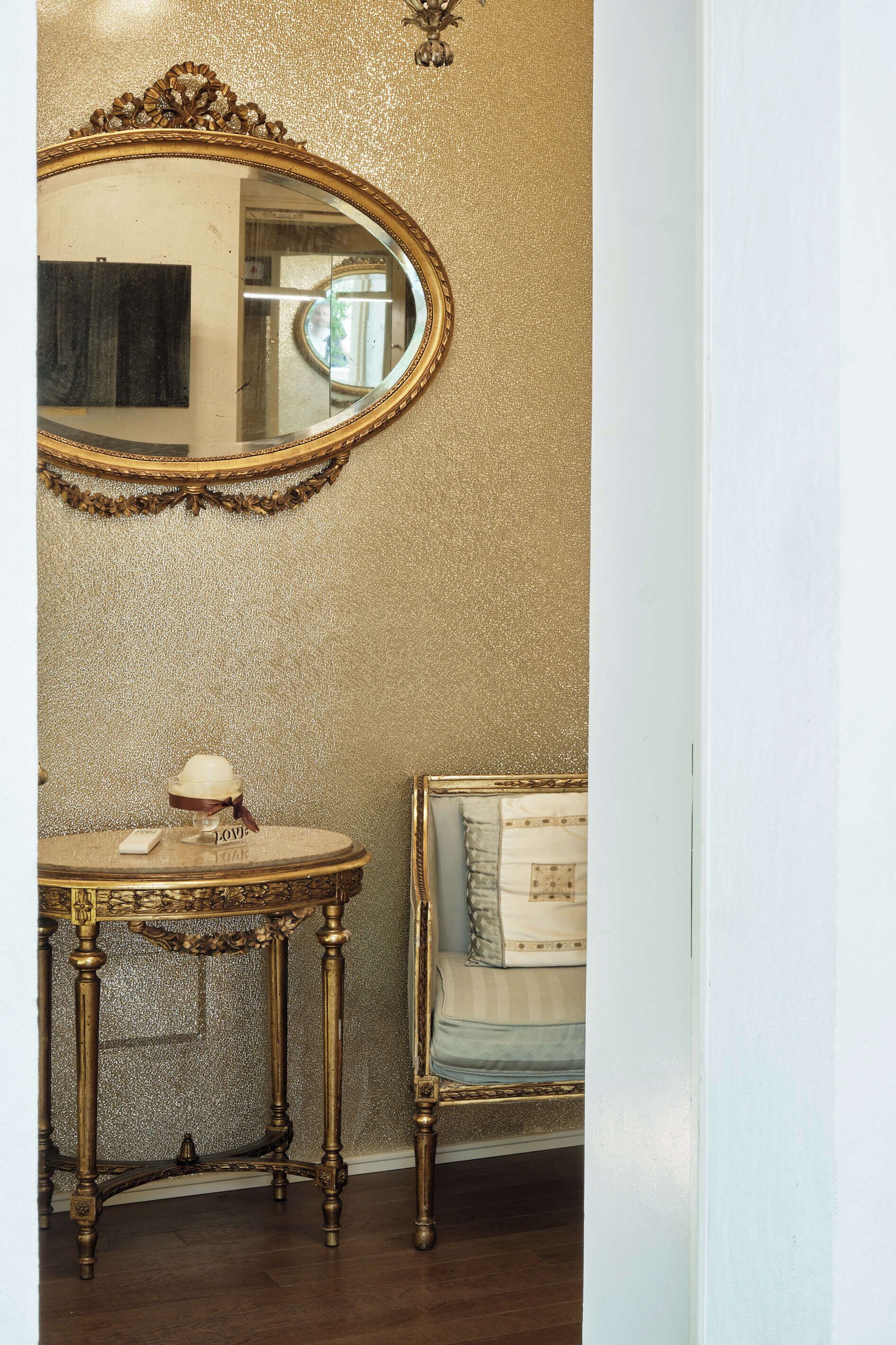 A room with a gold glittery wall, a round table with a cream-colored top, a cream-colored chair with a patterned cushion, and a large oval mirror with a gold frame.
