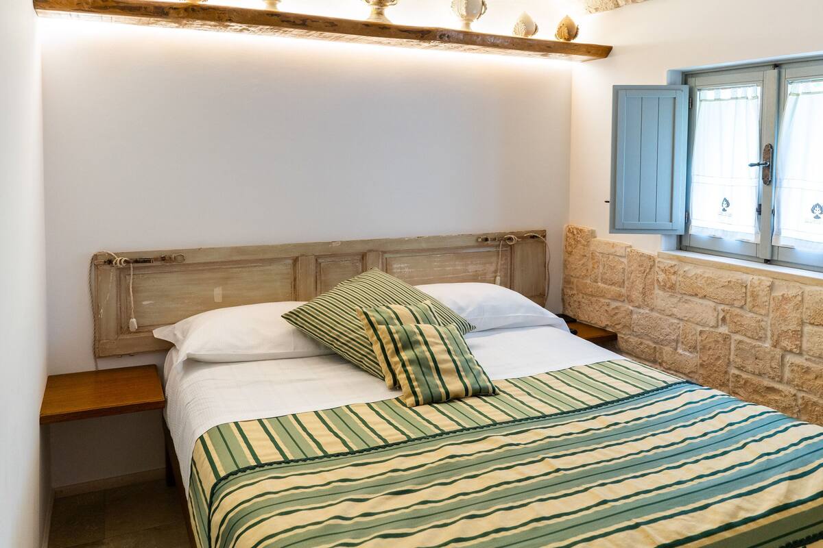 Une chambre avec un plafond en pierre, une tête de lit en bois, un lit avec une literie à rayures, une table de chevet et une fenêtre avec des volets bleus.