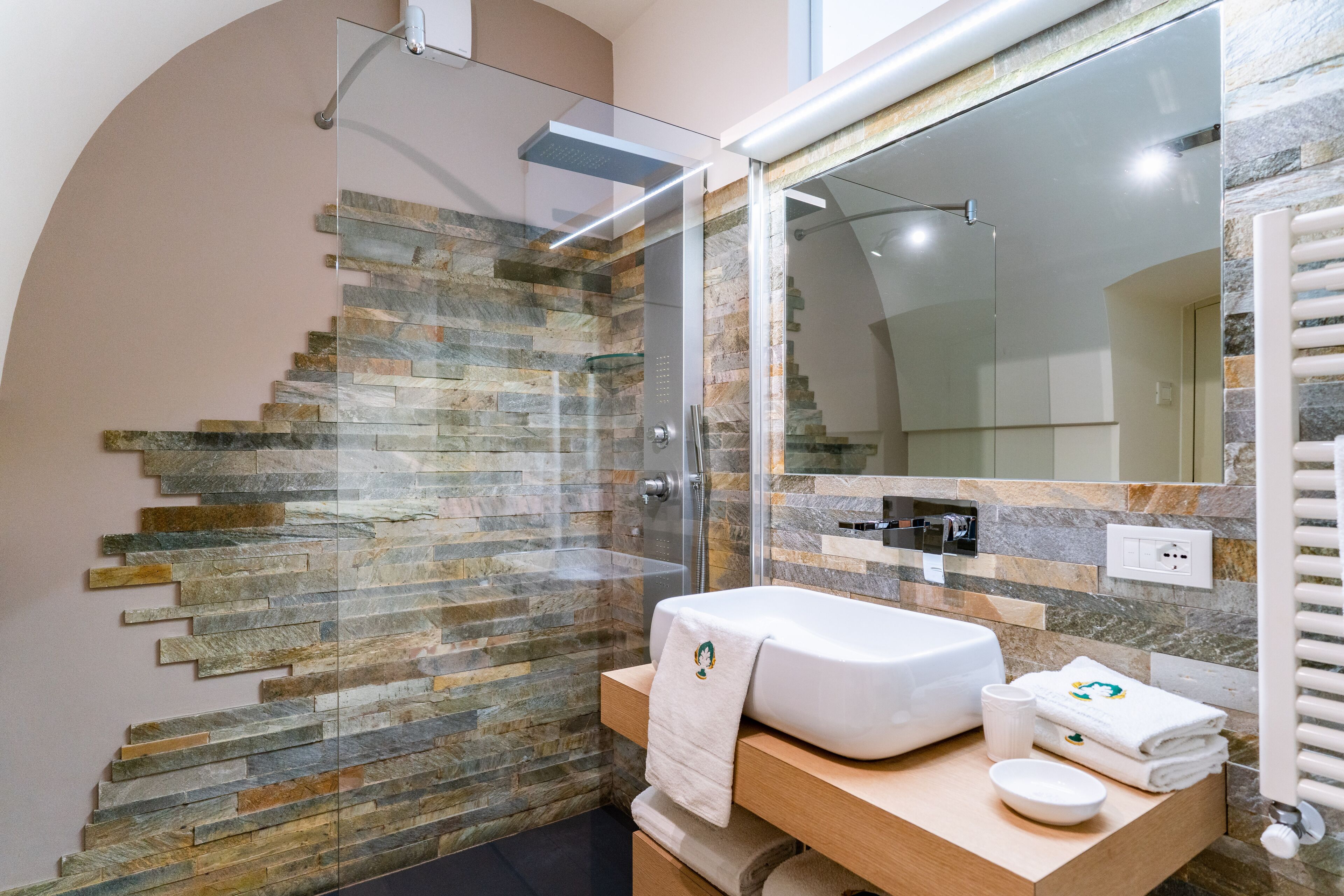 A modern bathroom with a glass-enclosed shower, a stone mosaic wall, a white sink, and a mirror above it.