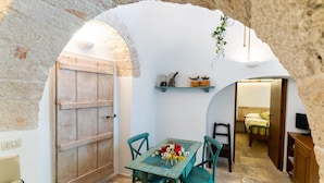A rustic room with a wooden door, teal chairs, a small table with flowers, and a bed in the background. - Palazzo Scotto (Alberobello)