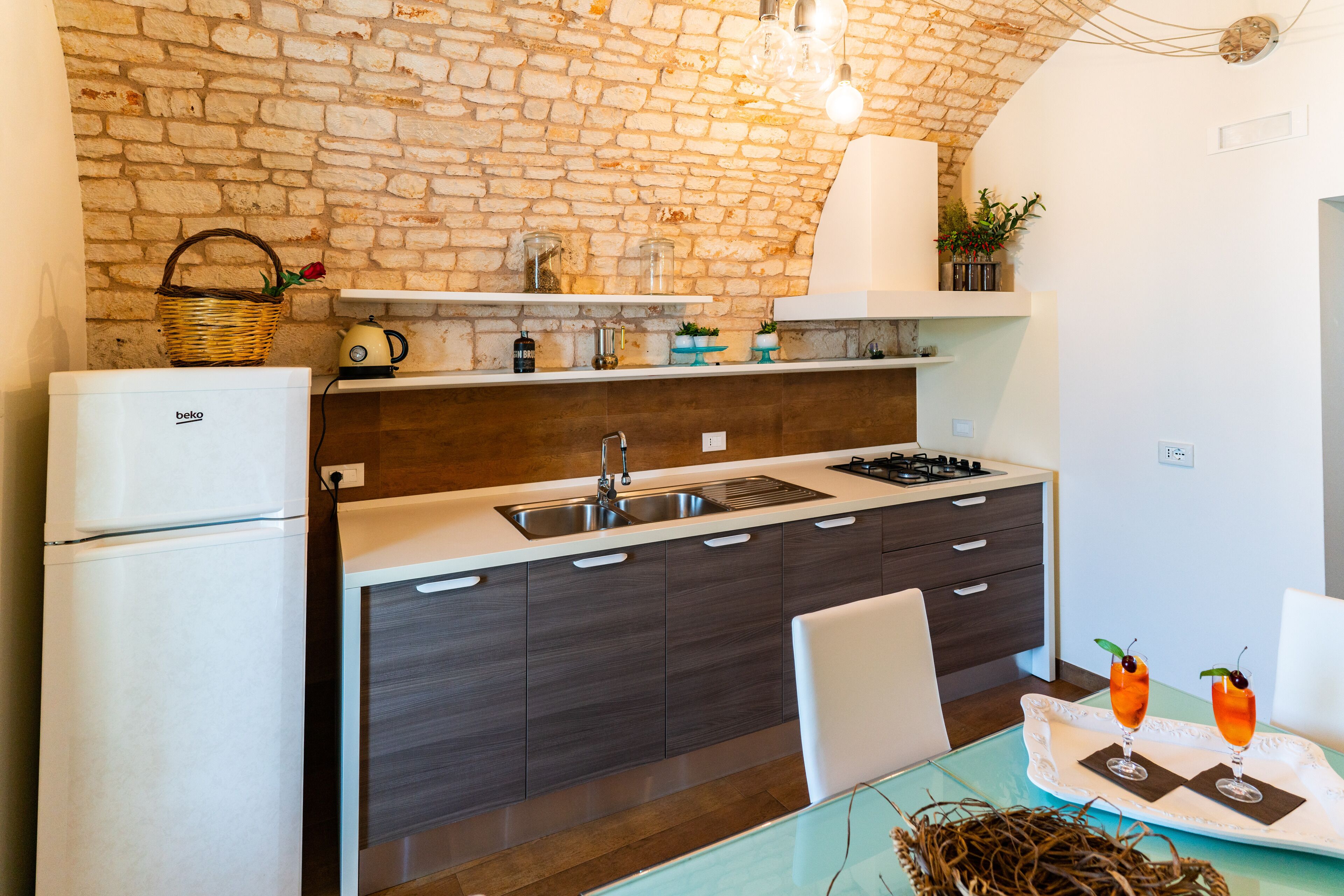 A modern kitchen with a brick wall, a refrigerator, a sink, and a dining area with a table and chairs.