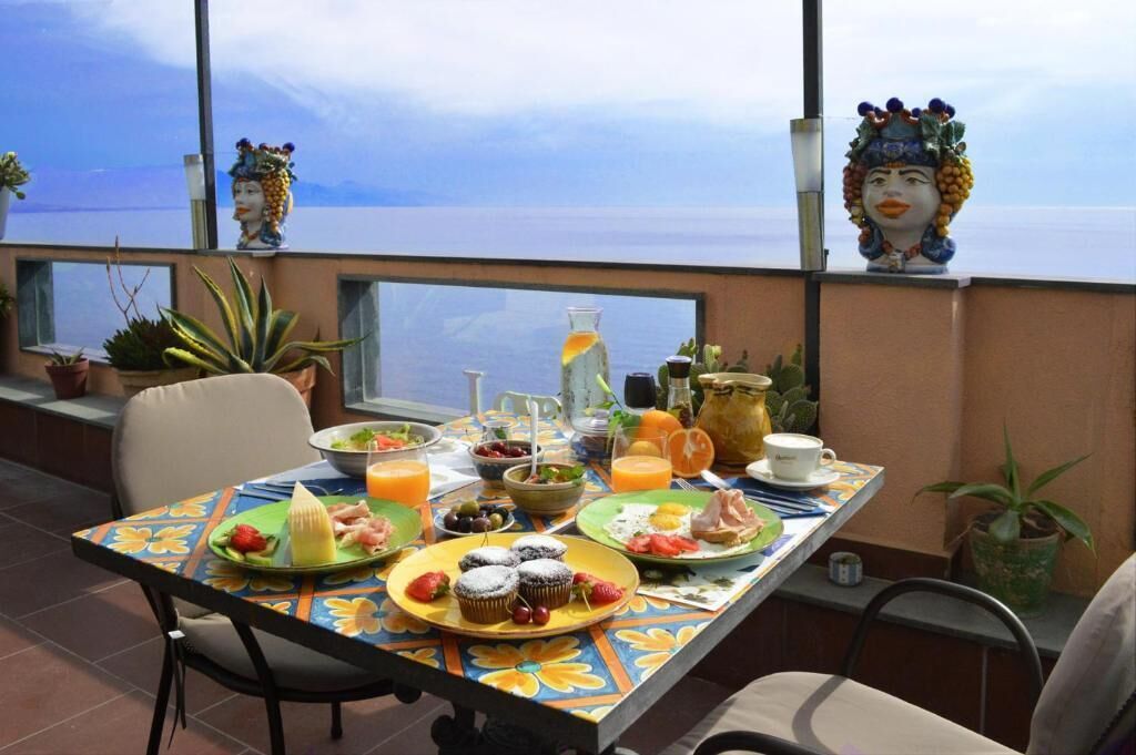 Mesa de comedor con alimentos para el desayuno, plantas en macetas y objetos decorativos en un balcón con vistas a una masa de agua.