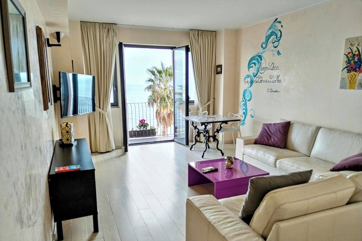 A modern living room with a sofa, a coffee table, a TV, and a balcony with a view of the sea.