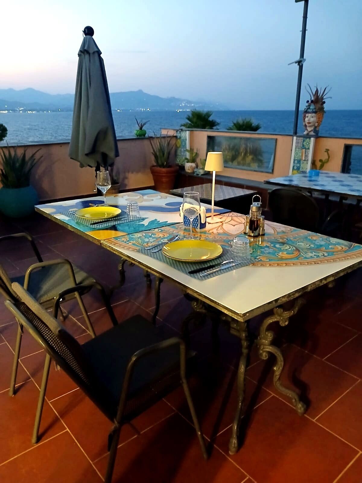 An outdoor dining area with a table set for two, chairs, a lamp, and a view of the sea.
