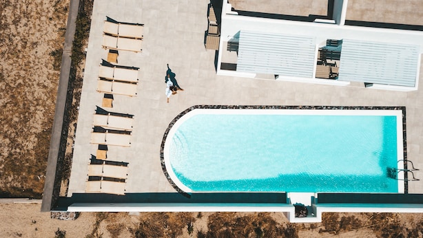 An aerial view of a swimming pool area with lounge chairs and a person walking. - Daylight (Santorini)