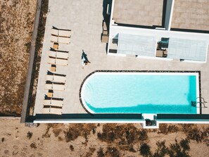 An aerial view of a swimming pool area with lounge chairs and a person walking. - Daylight (Santorini)