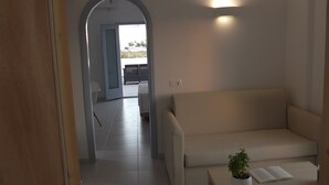 A living room with a beige sofa, a small table with a plant, and a doorway leading to another room. - Daylight (Santorini)