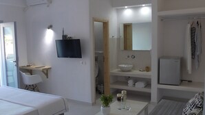 A hotel room with a bed, a TV mounted on the wall, a bathroom with a sink and mirror, and a small refrigerator. - Daylight (Santorini)