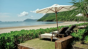 Een terras aan het strand met een houten bank en een grote parasol, omgeven door weelderige begroeiing en met uitzicht op zee.