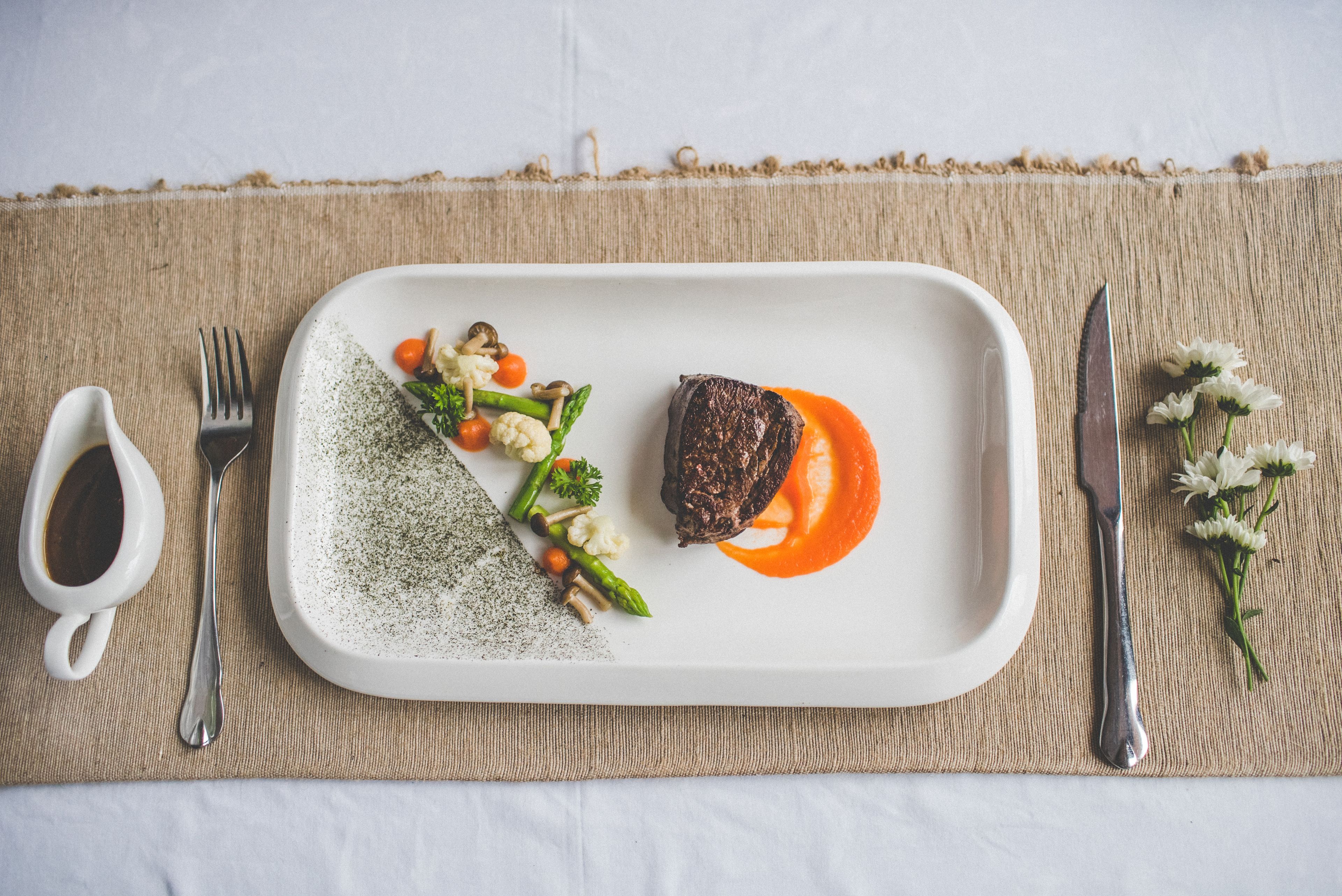 A rectangular white plate with a steak, vegetables, and sauce on a textured tablecloth.