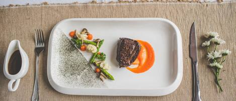 A rectangular white plate with a steak, vegetables, and sauce on a textured tablecloth.
