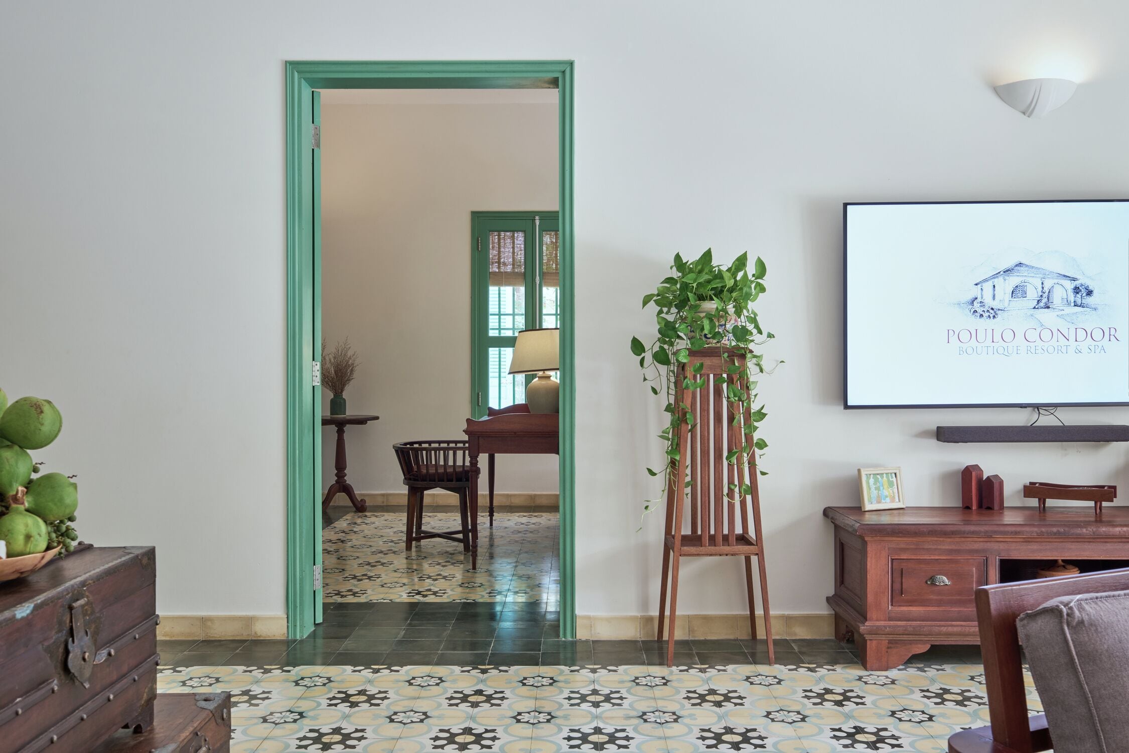 A room with a green-framed doorway leading to another room, a wooden dresser with a plant, a framed picture on the wall, and a tiled floor.