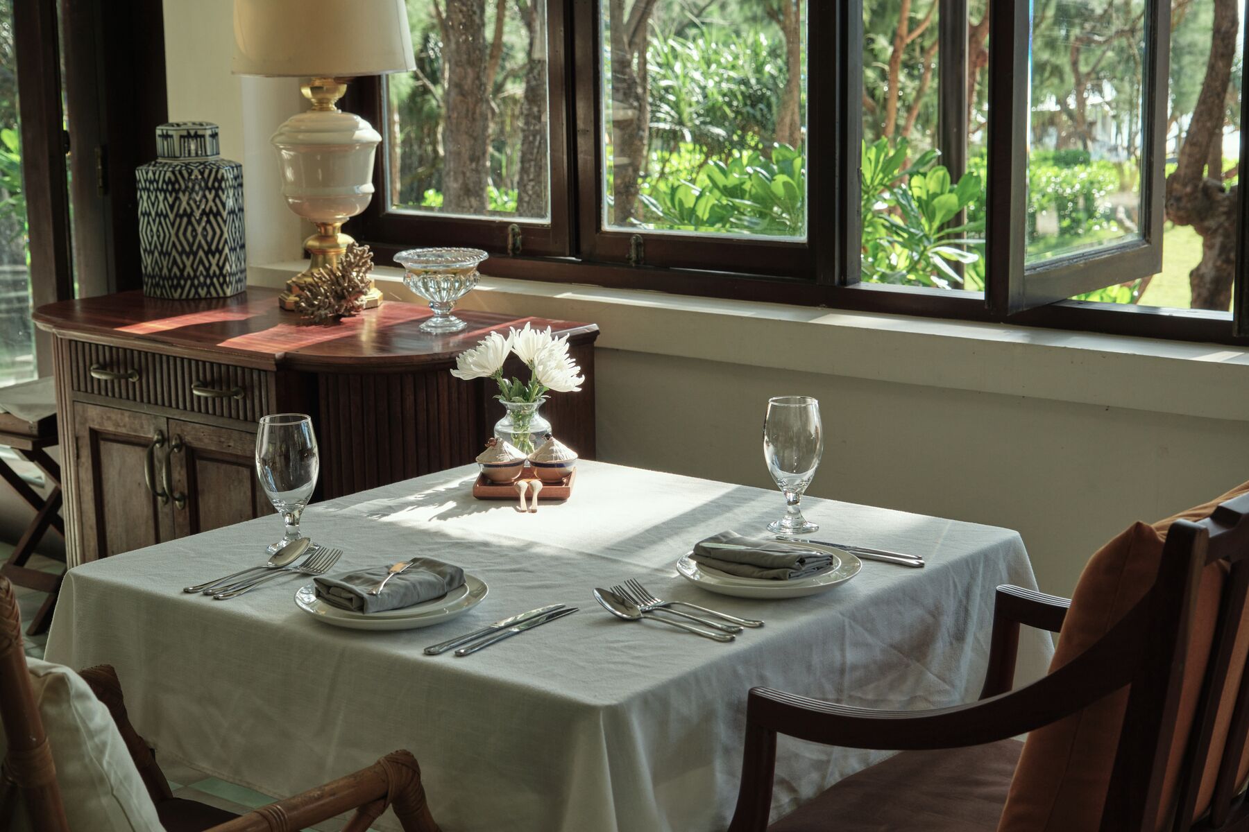 Una mesa de comedor lista con platos, cubiertos y vasos, con vistas a la vegetación por la ventana.