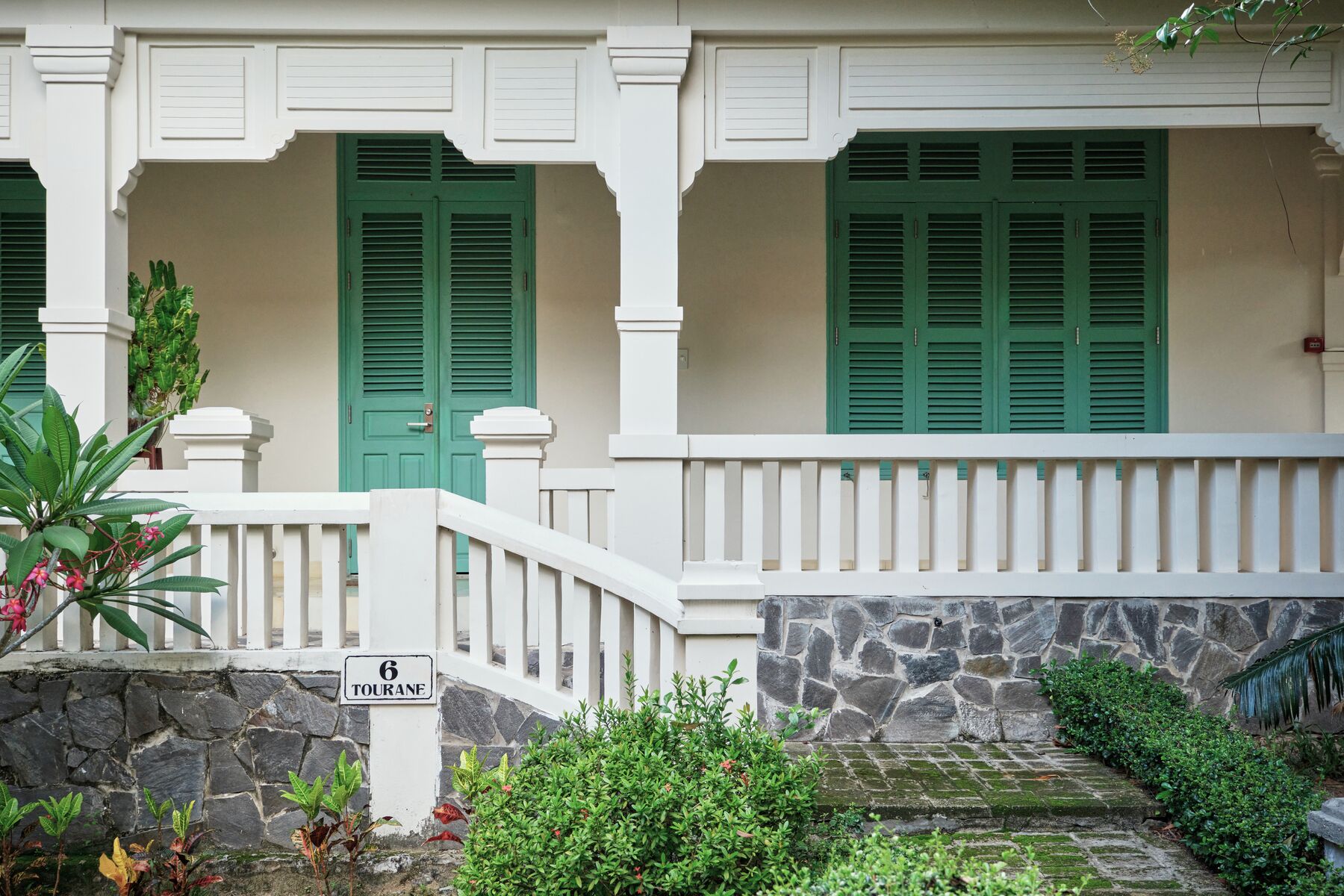A building with a stone wall, a sign reading "6 TOURANE", and green shutters on the windows.