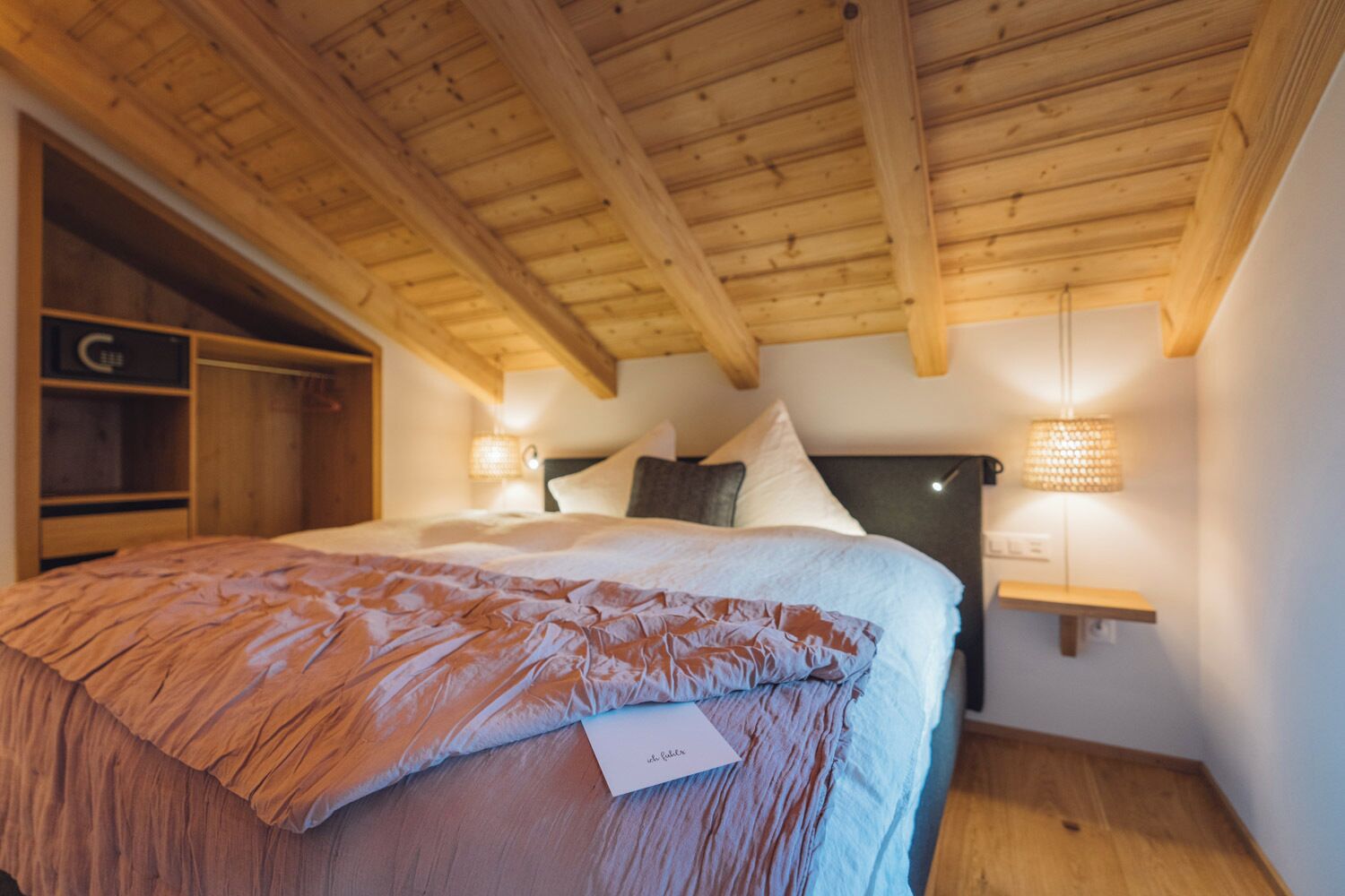 A bedroom with a wooden ceiling, a bed with a brown blanket, a nightstand with a lamp, and a wooden wardrobe.