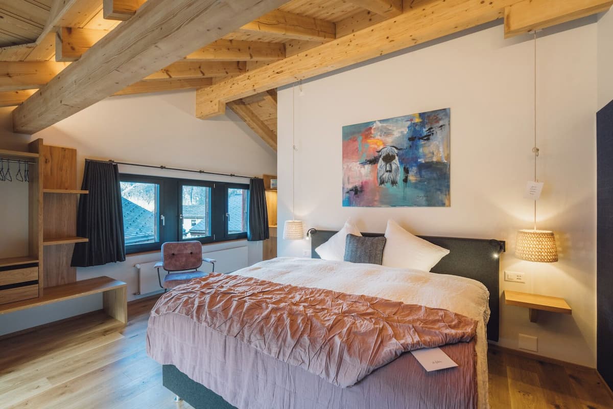 A bedroom with a large bed, wooden beams, a window, a chair, and a painting on the wall.