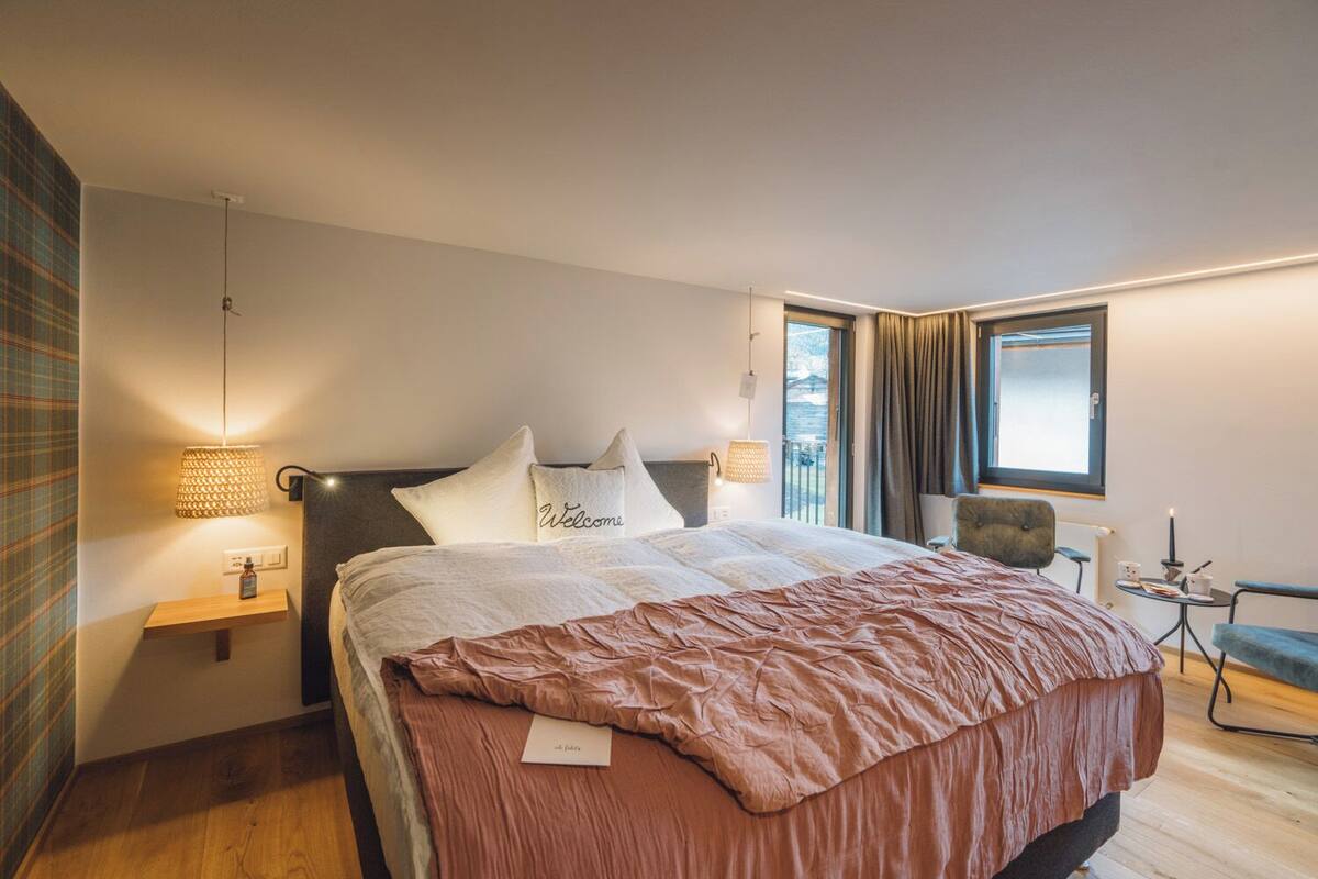 A modern hotel room with a large bed, a bedside table, a chair, a small table with a candle, and a window with curtains.