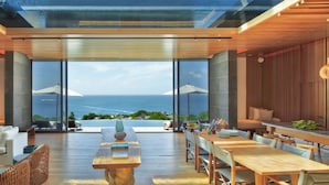 Un interior moderno con un amplio ventanal que ofrece vistas al océano. Hay una zona de estar con sofá y sillones, una mesa de comedor y una pequeña mesa decorativa con un libro.