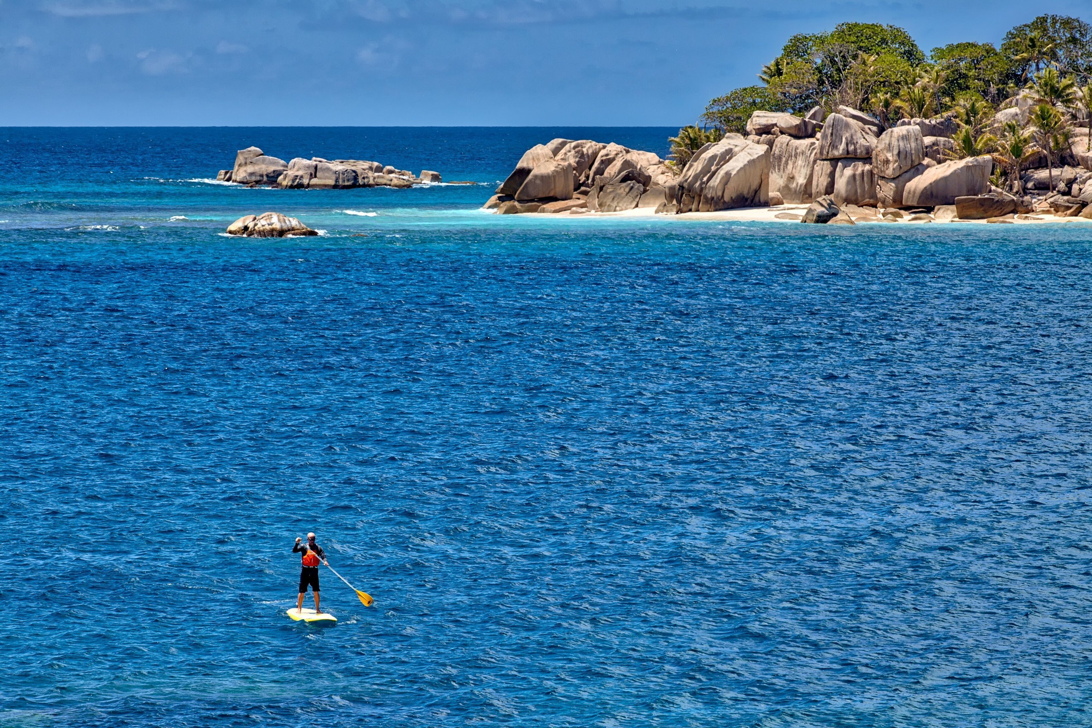 A person paddleboarding on a clear blue sea with rocky formations and palm trees in the background.
