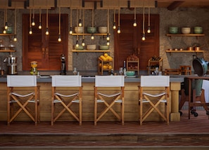 A rustic kitchen with wooden bar stools, hanging light bulbs, and shelves with bowls and fruits.