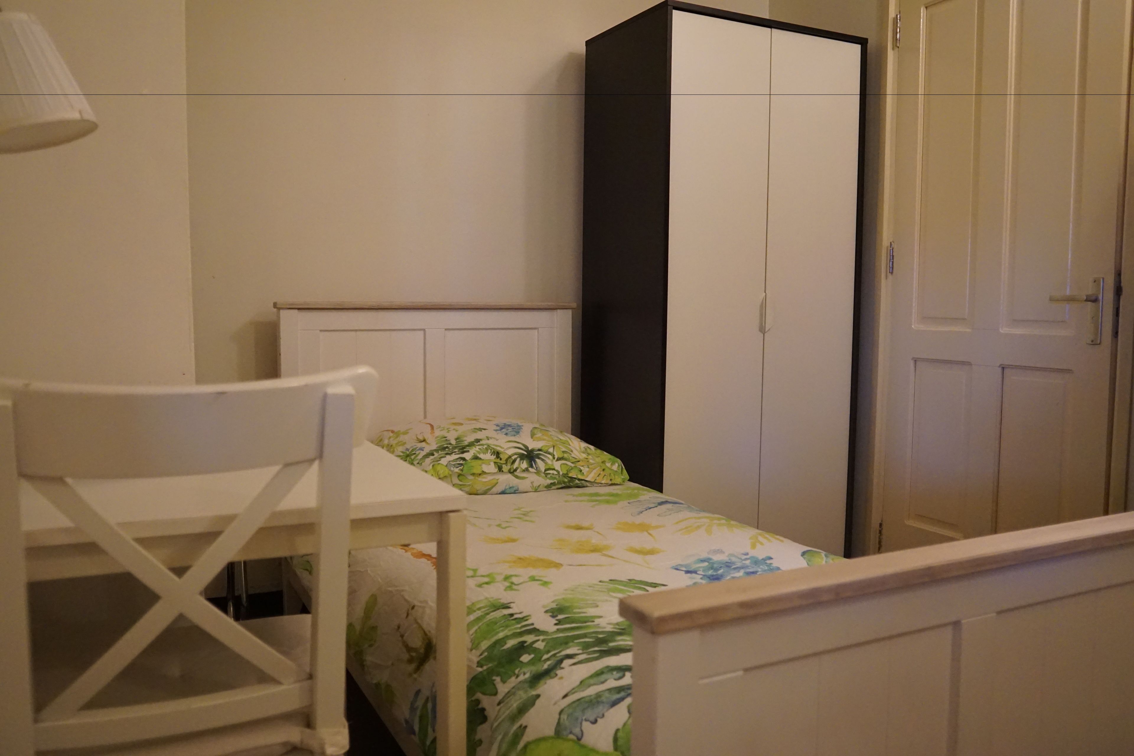 A small, neatly arranged bedroom with a single bed, a chair, a wardrobe, and a lamp.