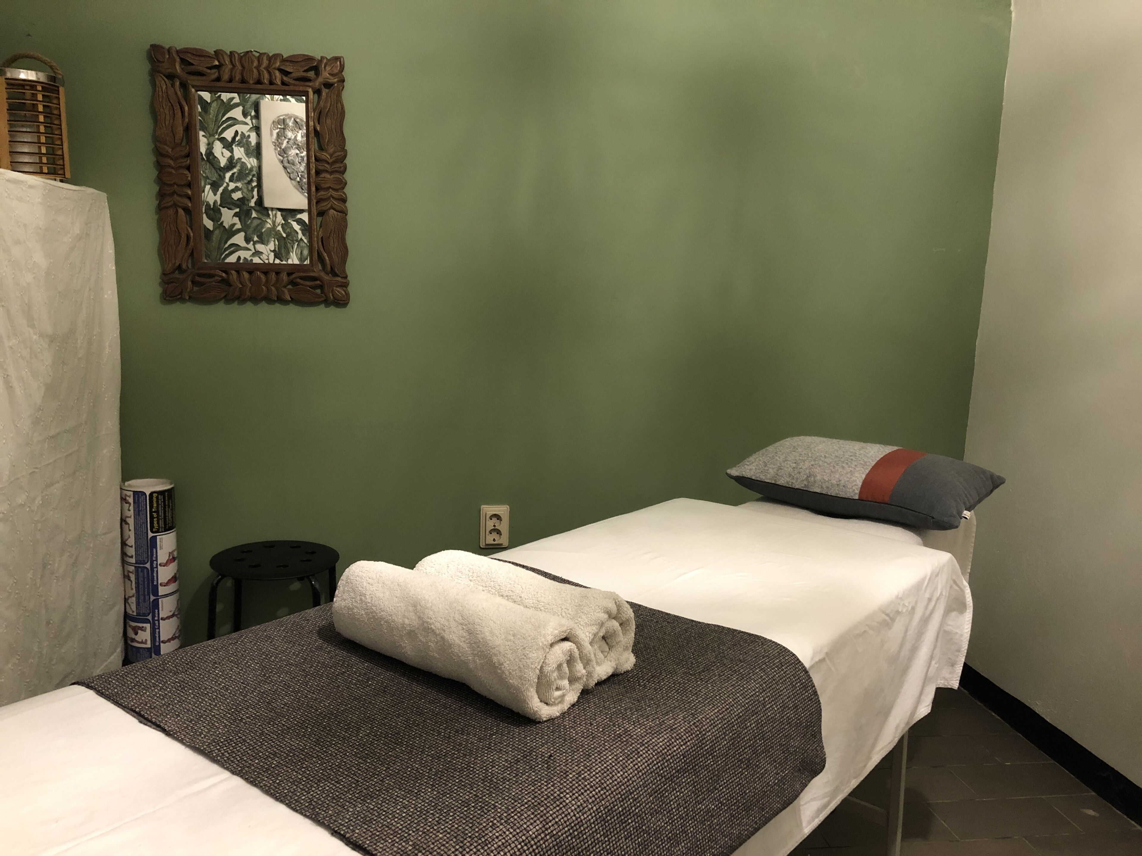 Una cama individual con una colcha blanca, una toalla enrollada, un almohadón gris y un espejo decorativo sobre una pared verde.