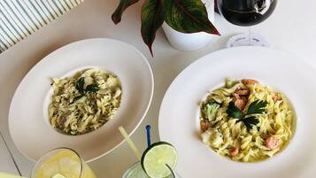Two white plates of pasta with garnishes, a glass of orange juice, a glass of iced drink with lime, and a glass of red wine on a table.