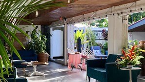 Un'ampia area interna con soffitto alto, travi in legno e lampade a sospensione. Diverse zone salotto con divani e poltrone, circondate da piante in vaso. Sullo sfondo, un tavolo con tovaglia rosa.