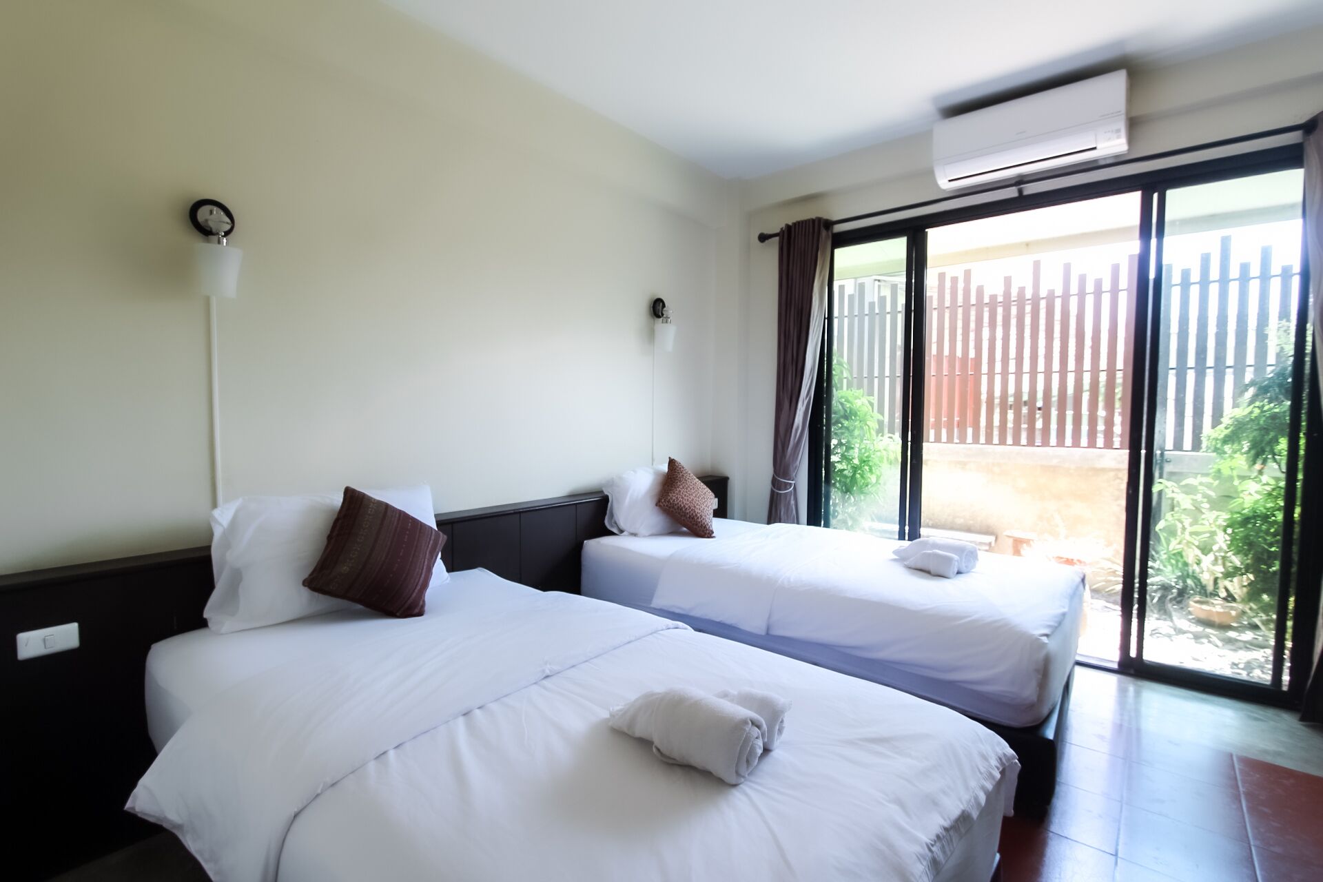 A hotel room with two beds, a large window with a view of a fence and greenery, and an air conditioning unit on the wall.
