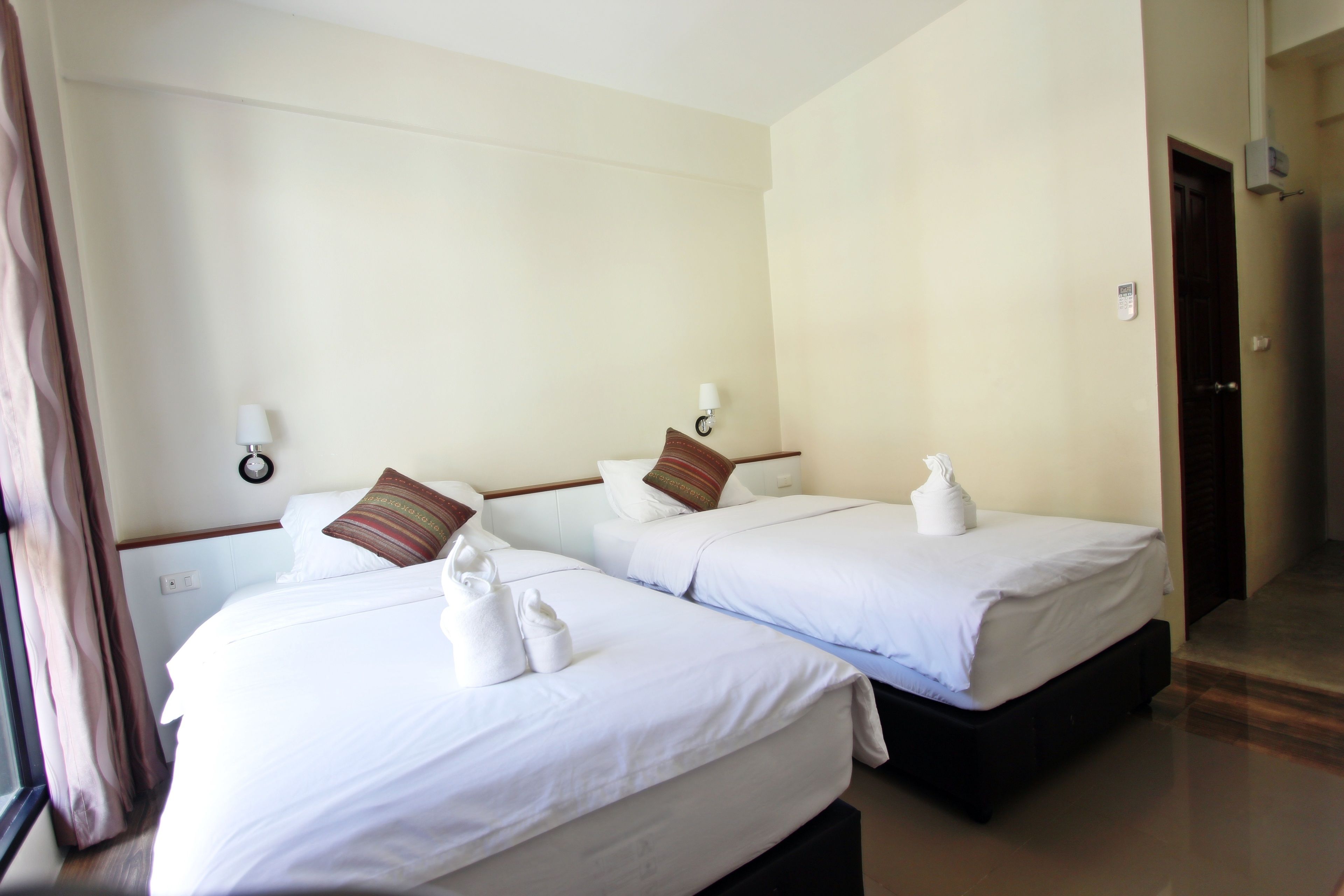 Habitación de hotel con dos camas, cada una con sábanas y almohadas blancas, una mesita de noche, una lámpara y una puerta.
