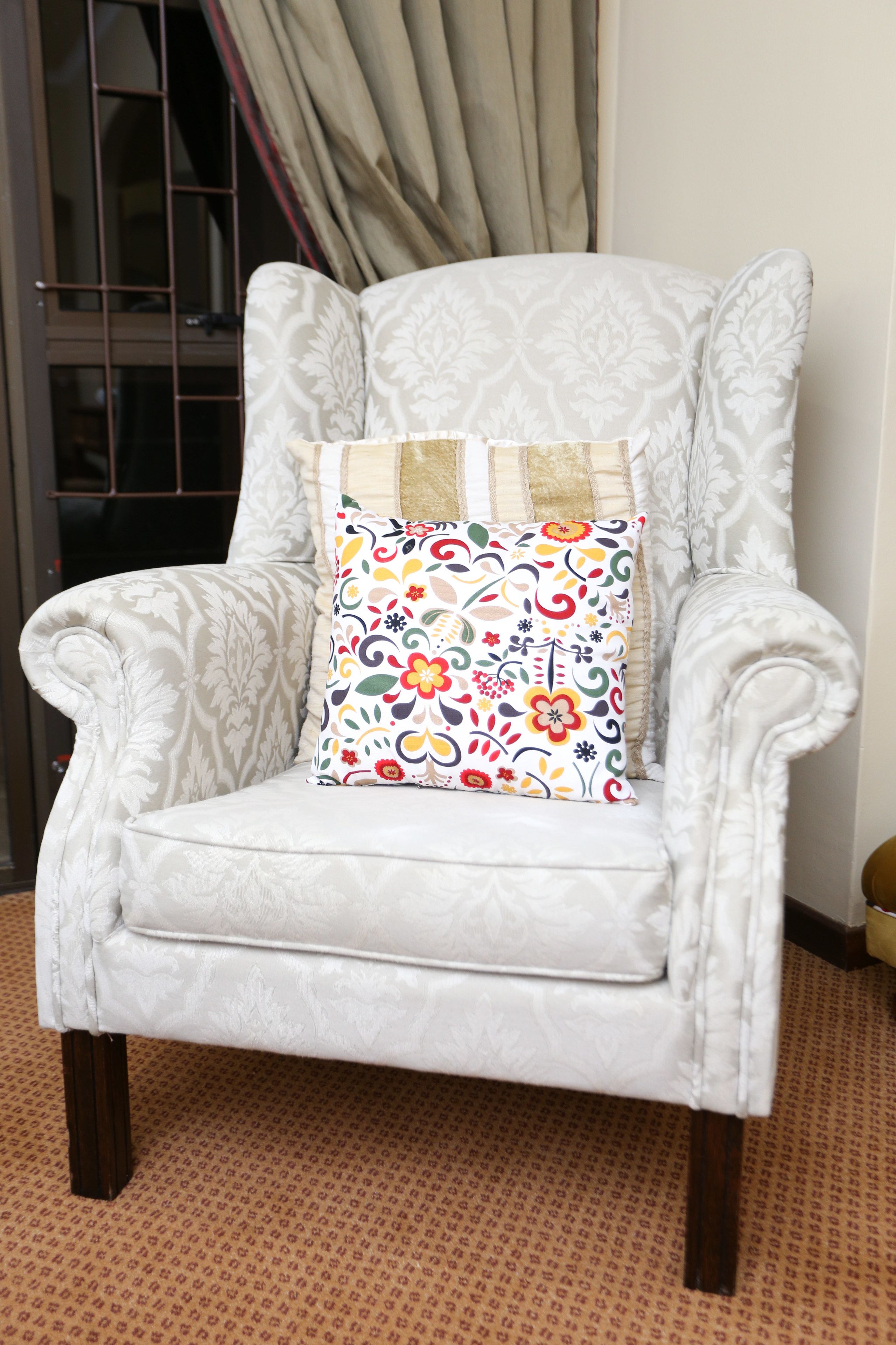 A white armchair with a floral cushion, positioned in a room with patterned carpet and a window with curtains.