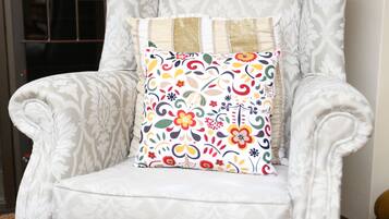 A white armchair with a floral cushion, positioned in a room with patterned carpet and a window with curtains.