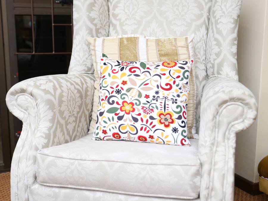 A white armchair with a floral cushion, positioned in a room with patterned carpet and a window with curtains.