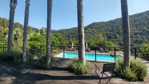 A wooden deck with a pool, surrounded by palm trees and a view of a forested hill.