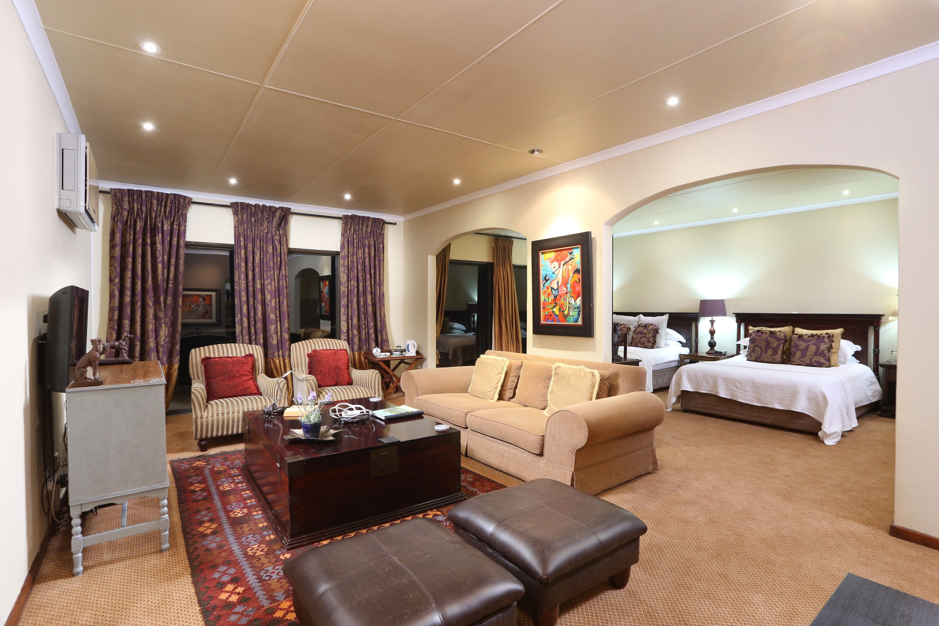 A hotel room with a living area, a sofa, a coffee table, an ottoman, a bed, and a TV.