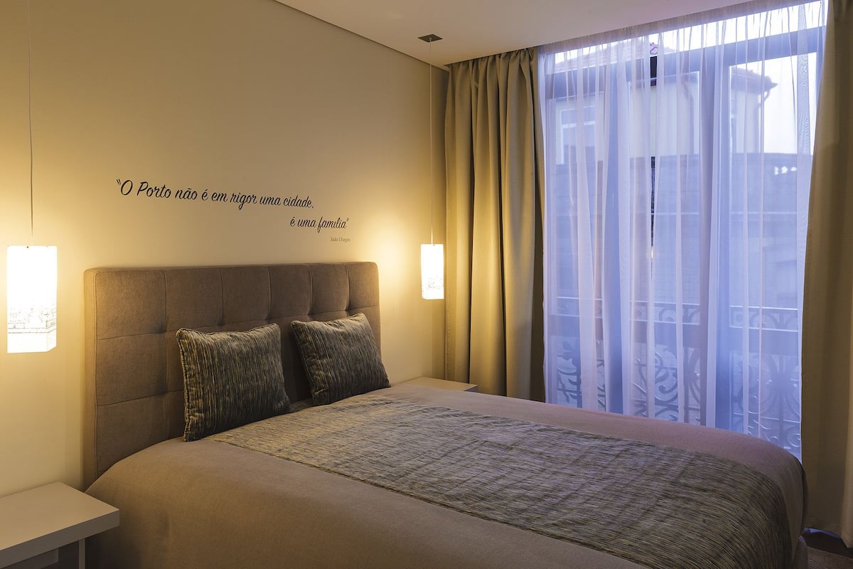 A hotel room with a large bed, two pillows, a headboard, a bedside table, a wall-mounted light, and a window with sheer curtains.
