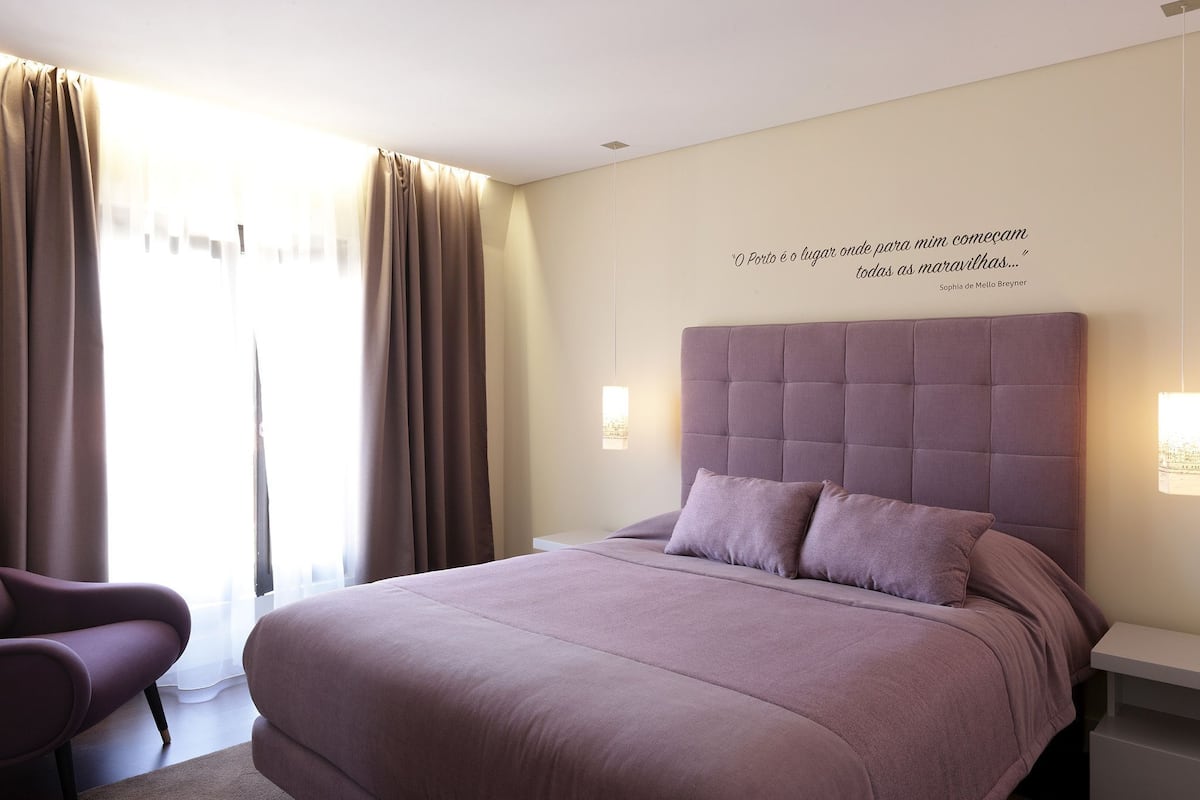 A bedroom with a large bed, a chair, a wall quote, and a window with curtains.