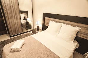 A neatly made bed with a rolled towel, a bedside table with a lamp, and a mirror reflecting part of the room.
