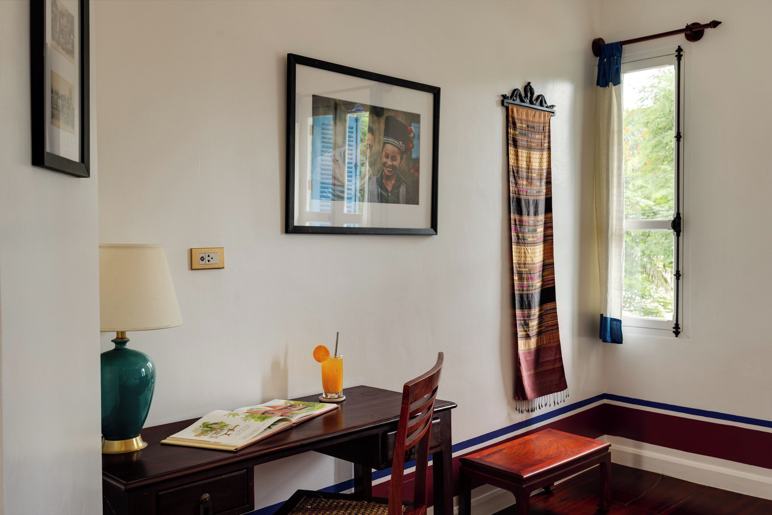 Een kamer met een bureau, stoel, lamp, ingelijst kunstwerk en een raam met gordijn.