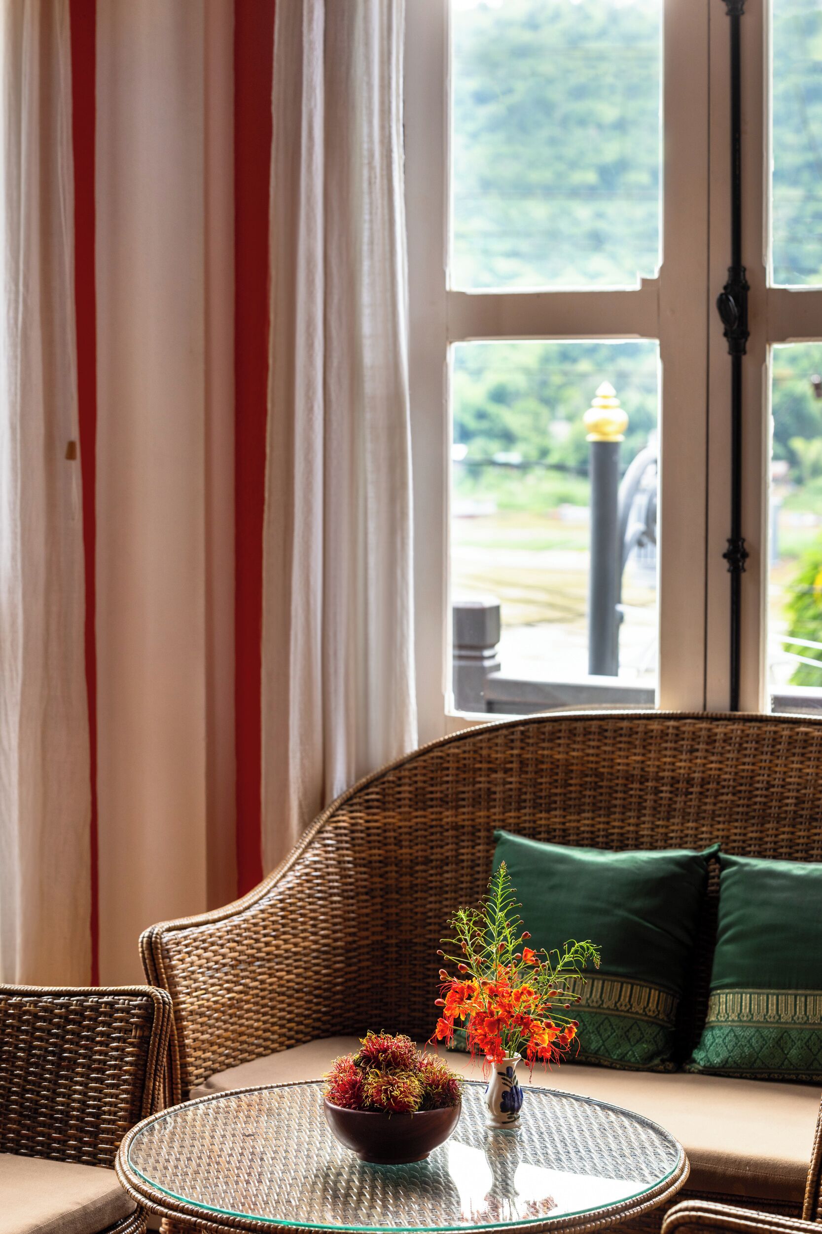 A wicker sofa with a glass table in front, a bowl of flowers, and a view of greenery outside.