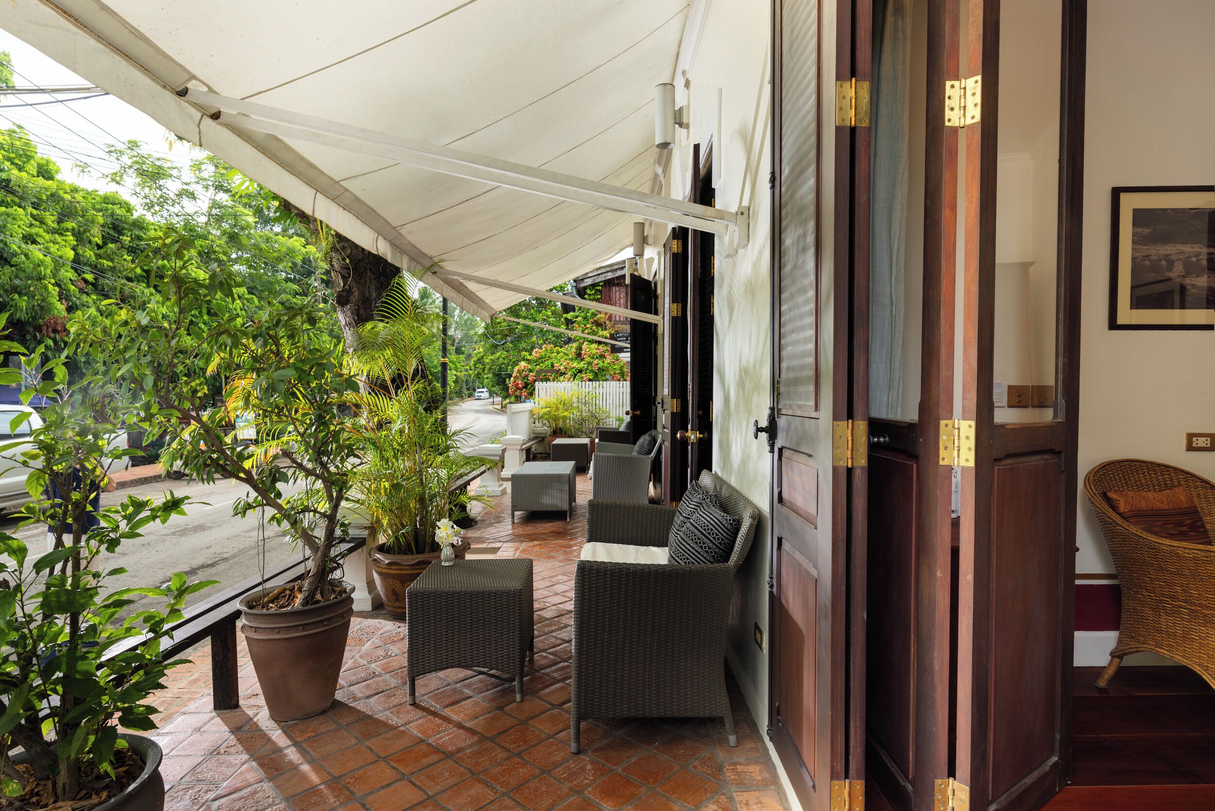 A covered outdoor seating area with potted plants, wicker furniture, and a view of the street.