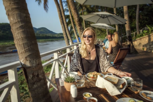 A dining table with food and drinks, a potted plant, and a view of a river and mountains. - The Belle Rive Boutique Hotel (Luang Prabang)