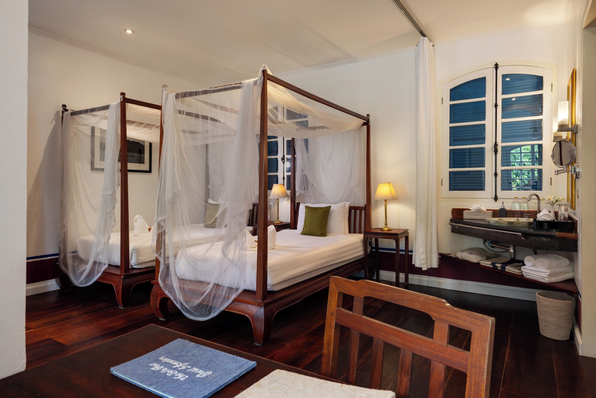 A four-poster bed with a canopy, a bedside table with a lamp, a mirror, and a window with blinds.