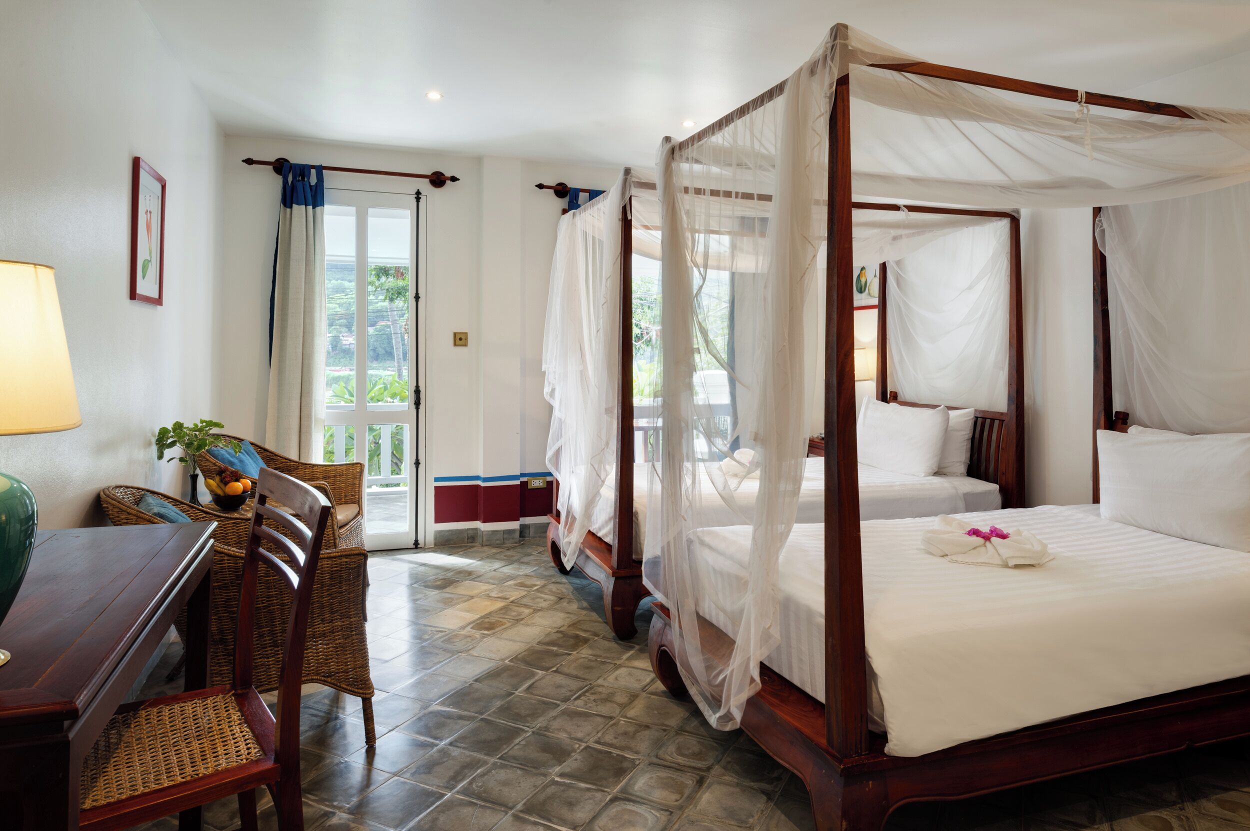 A four-poster bed with a canopy, a dining area with wicker furniture, and a view of greenery through the windows.
